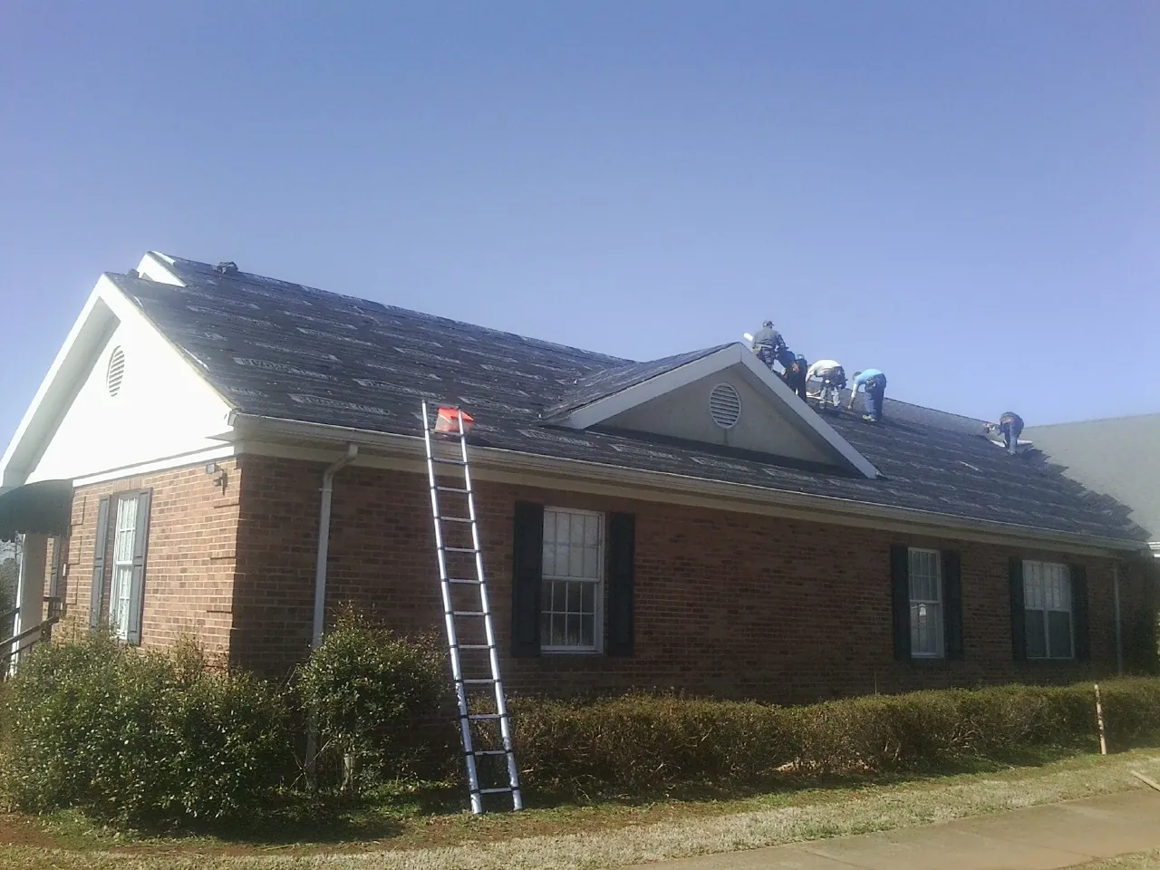 Crew tearing shingles off a brick ranch roof with a ladder set — Metal Roof Repair work by our crew in Roebuck