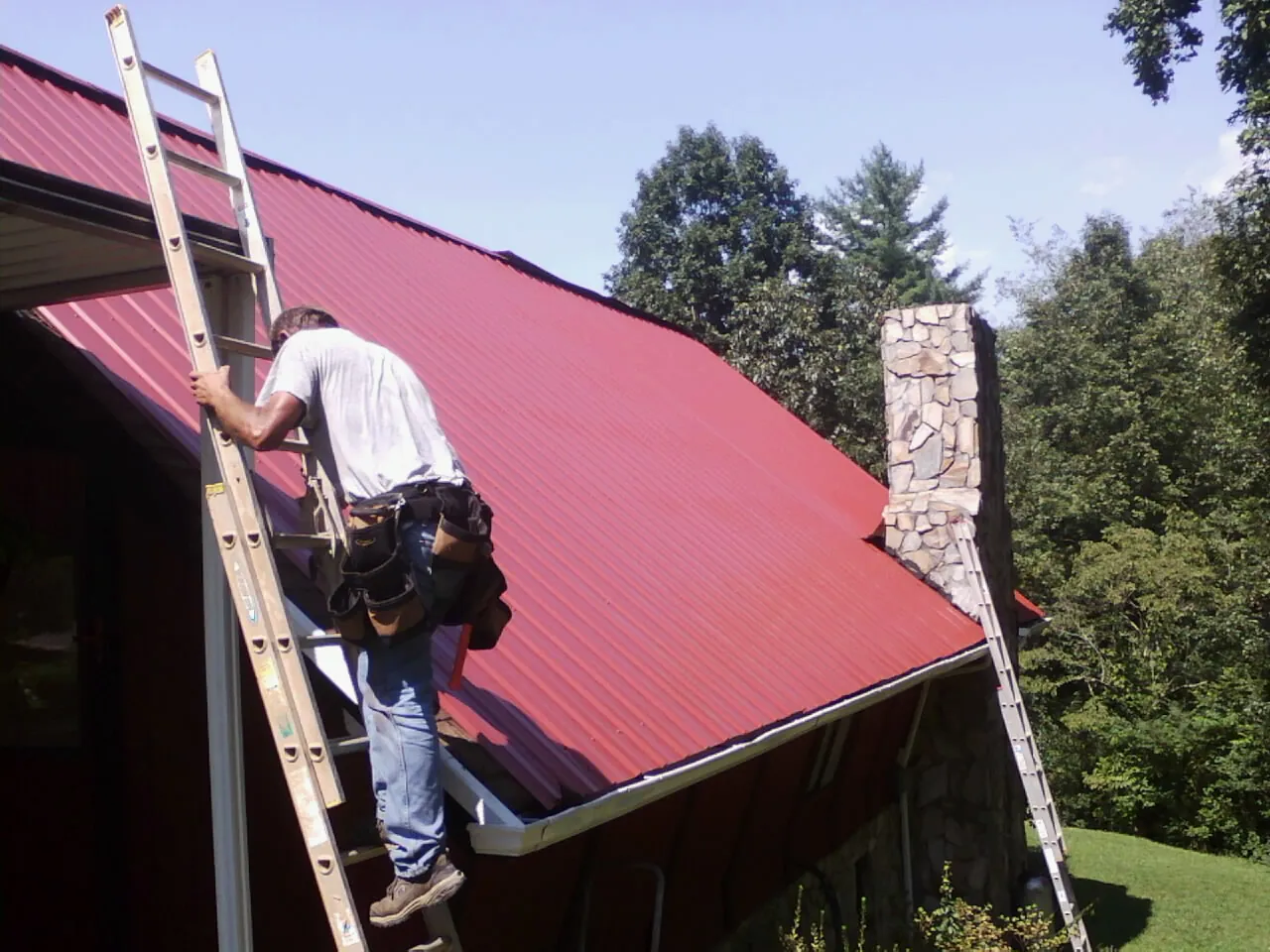 Roofer on a ladder beside a red metal roof and stone chimney — Soffit & Fascia Repair by Roofing Brothers in Roebuck